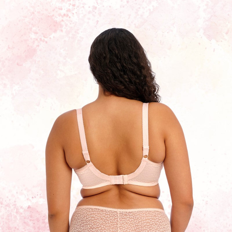En kvinde med medium hudtone og langt krøllet hår vender ansigtet væk fra kameraet. Hun er modelleret af en lyserød Elomi Lucie Stretch Bh og matchende undertøj med et diskret mønster mod en akvarelrosa baggrund.