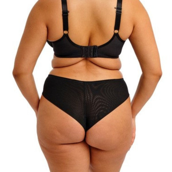 En person med en fyldigere figur ses bagfra iført en sort Elomi-bh og en brasiliansk Reja-trusse. Deres naturlige kurver fremhæver den halvdækkende pasform, med armene afslappet i siden, mod en almindelig hvid baggrund.
