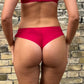En person vender ansigtet væk fra kameraet iført en Fantasie rød trusse bikini med broderidetaljer. Deres solbrune hud står i kontrast til den strukturerede, lyse murstensvæg. Billedet viser kun den nederste del af ryggen, taljen og den øverste del af benene.