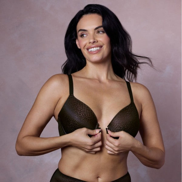En kvinde med langt mørkt hår smiler, mens hun spænder en mørkegrøn Wacoal bh med leopardprint foran. Hun står på en lyslilla baggrund, kigger lidt til siden og justerer bh'en med begge hænder.