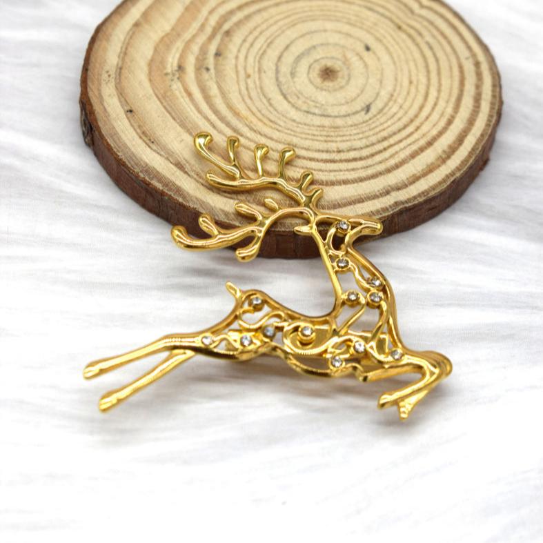 En guldfarvet, udsmykket Rouge Bonbons-broche med et gennembrudt rensdyrmotiv og funklende sten hviler foran en rund træplade på en blød, hvid struktureret overflade.